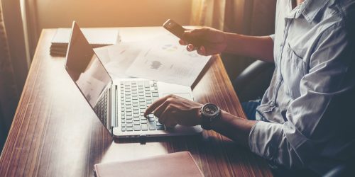 businessman-hands-using-cell-phone-with-laptop-at-office-desk